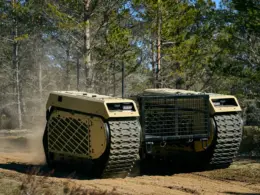 A Milrem THeMIS unmanned ground vehicle crosses sandy terrain in a forest, raising dust behind its tracks.