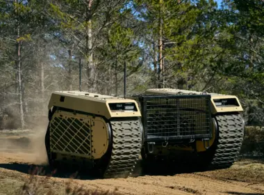 A Milrem THeMIS unmanned ground vehicle crosses sandy terrain in a forest, raising dust behind its tracks.