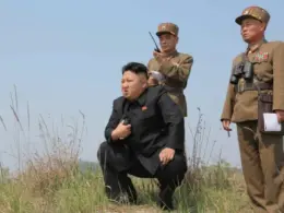 North Korean leader Kim Jong-un crouches in a grassy field while two uniformed military officers stand behind him.