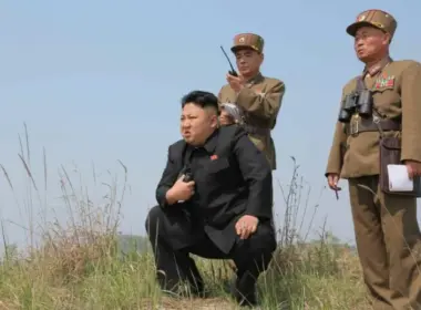 North Korean leader Kim Jong-un crouches in a grassy field while two uniformed military officers stand behind him.