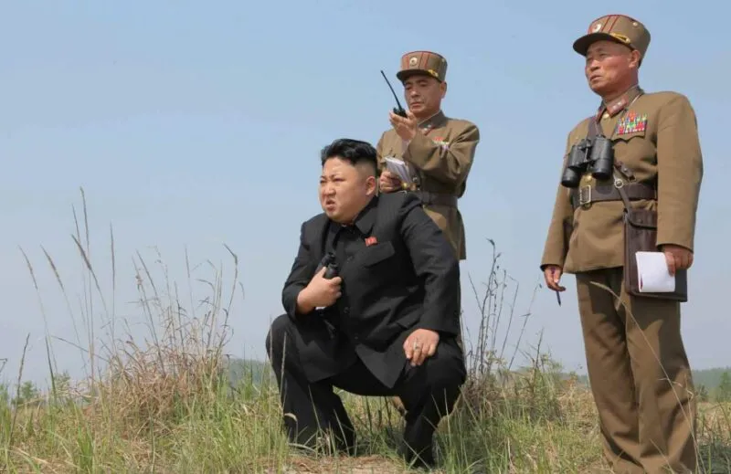 North Korean leader Kim Jong-un crouches in a grassy field while two uniformed military officers stand behind him.