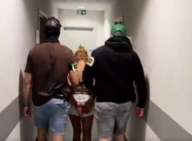 A woman with her hands behind her back walks between two masked police officers down a white corridor.