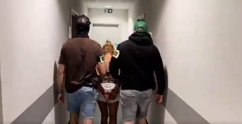 A woman with her hands behind her back walks between two masked police officers down a white corridor.