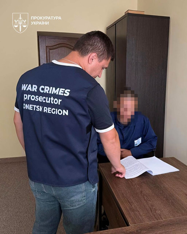 A man wearing a vest marked “War Crimes Prosecutor, Donetsk Region” stands beside a seated man with a pixelated face as they look at documents on a desk.