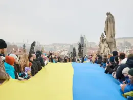 People line Prague’s Charles Bridge holding a long Ukrainian flag, with Prague Castle visible in the background.