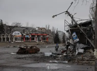 A person walks along a war-damaged street past a burned-out car, rubble, shattered buildings and a collapsed utility pole.