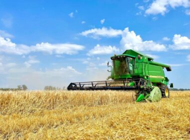 A green combine harvester cuts wheat beneath a blue, partly cloudy sky.