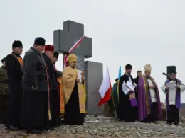 Polish and Ukrainian clergy stand beside a memorial cross during a ceremony, with both countries’ flags behind them.