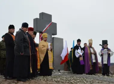 Polish and Ukrainian clergy stand beside a memorial cross during a ceremony, with both countries’ flags behind them.