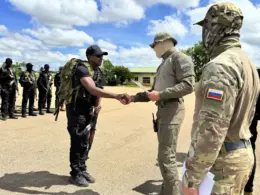 A soldier in a black uniform shakes hands with a masked man holding papers as another uniformed man with a Russian flag patch looks on.