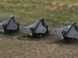 Three gray, triangular-wing drones sit on camouflage netting in a grassy field.