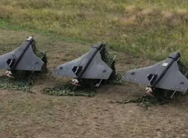 Three gray, triangular-wing drones sit on camouflage netting in a grassy field.