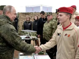 Vladimir Putin shakes hands with a young Yunarmia member wearing a beige uniform and red beret as other members stand behind them.