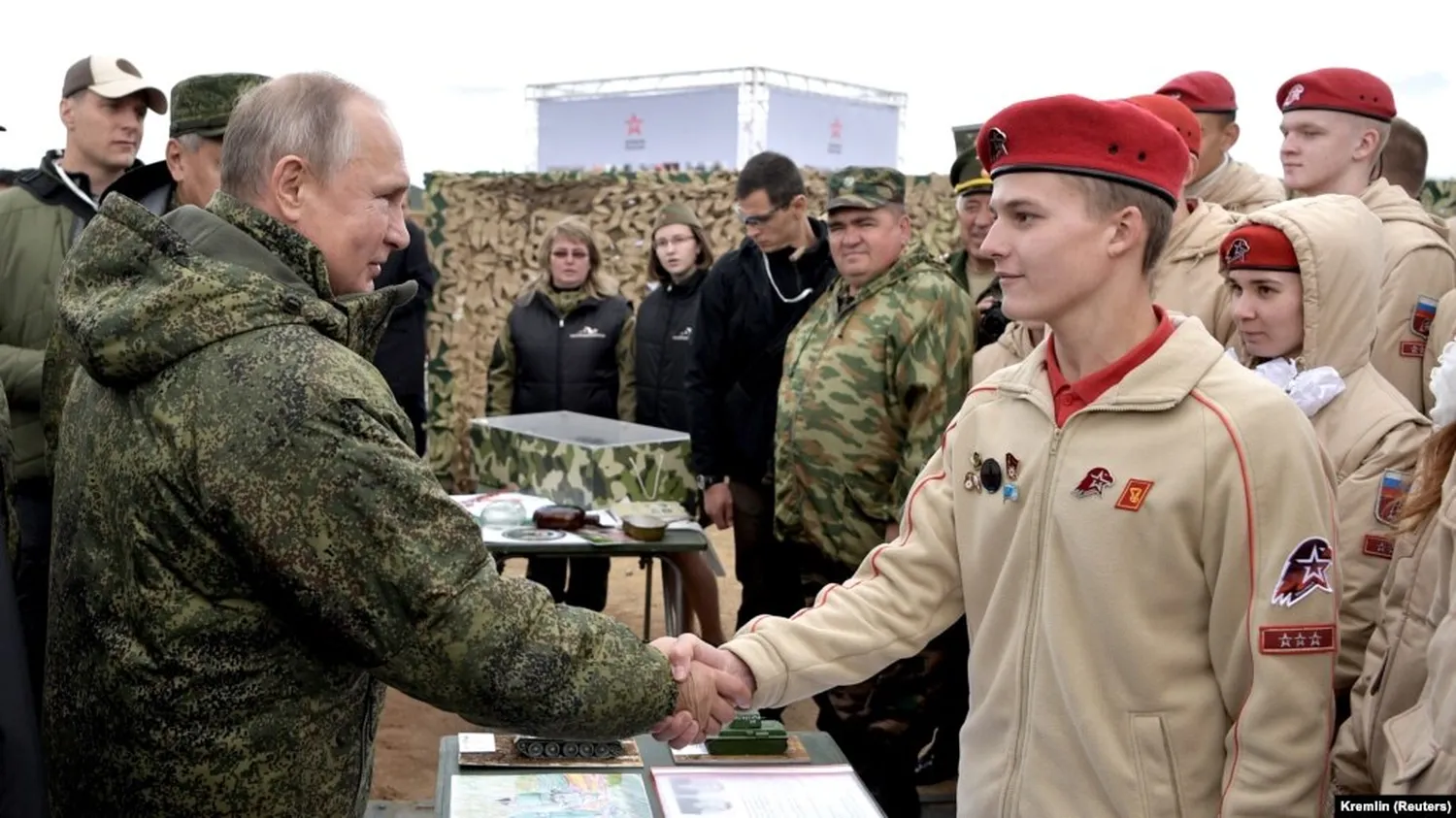 Vladimir Putin shakes hands with a young Yunarmia member wearing a beige uniform and red beret as other members stand behind them.