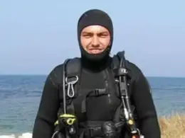 Volodymyr Zhuravliov wears a black hooded wetsuit and scuba harness while standing beside the sea.
