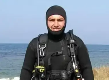 Volodymyr Zhuravliov wears a black hooded wetsuit and scuba harness while standing beside the sea.