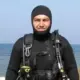 Volodymyr Zhuravliov wears a black hooded wetsuit and scuba harness while standing beside the sea.