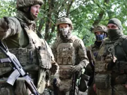 A unit commander gestures while briefing Colombian and Ukrainian soldiers wearing camouflage and tactical gear in a wooded area.