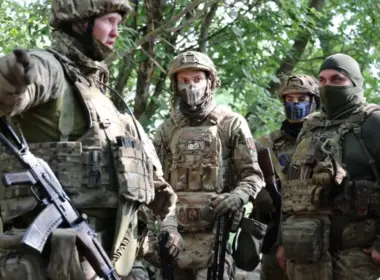 A unit commander gestures while briefing Colombian and Ukrainian soldiers wearing camouflage and tactical gear in a wooded area.