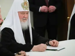Patriarch Kirill sits at a white table in black-and-white Orthodox vestments, looking toward the camera as several people stand around him.
