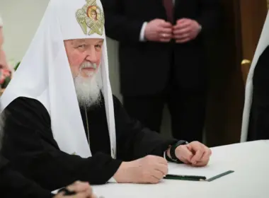 Patriarch Kirill sits at a white table in black-and-white Orthodox vestments, looking toward the camera as several people stand around him.