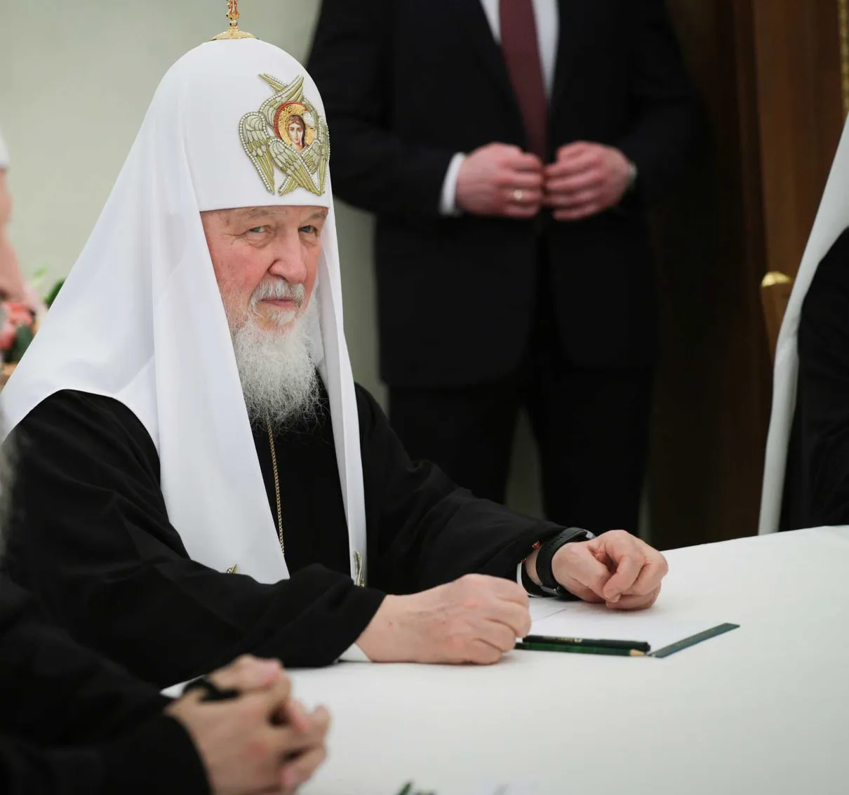 Patriarch Kirill sits at a white table in black-and-white Orthodox vestments, looking toward the camera as several people stand around him.