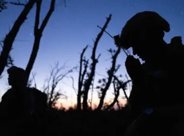 Two soldiers of Ukraine’s Third Army Corps silhouetted among bare trees at twilight; one holds a radio to his mouth.