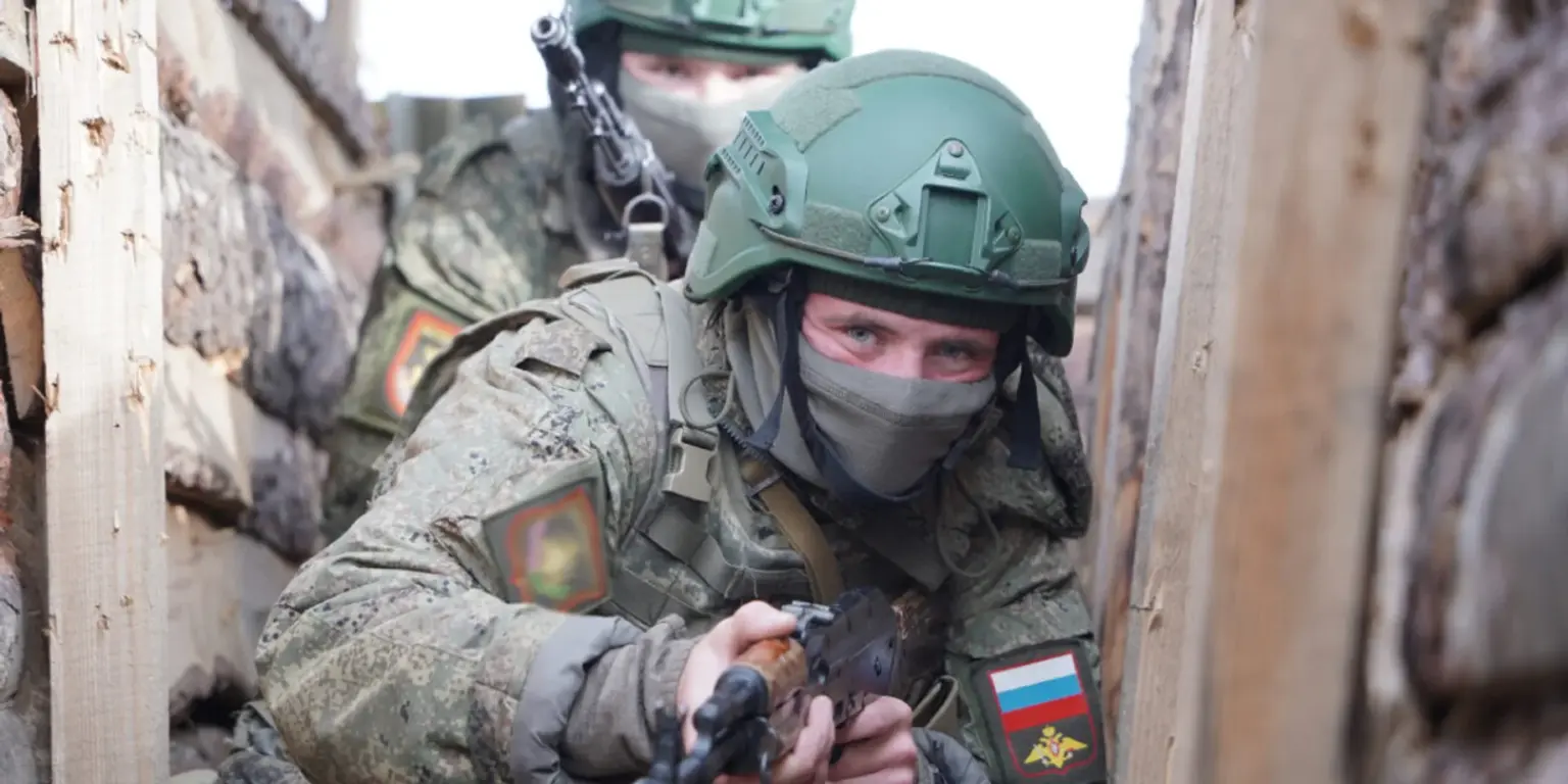 Two masked soldiers in camouflage crouch in a narrow trench. The soldier in front holds a rifle and wears a Russian flag patch.