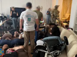SBU and Prosecutor General’s Office personnel stand inside a crowded room with computers as several people with pixelated faces lie or sit on the floor. An armed tactical officer stands near the back.