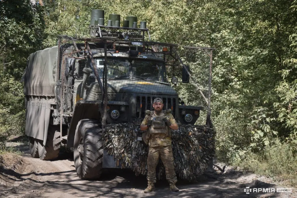 A soldier with the call sign Tatarin in front of a truck fitted with anti-drone netting and camouflage.