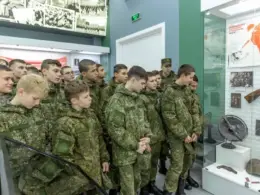 Children and teenagers in matching green camouflage stand beside a museum display of rifles, helmets, and historical photographs.