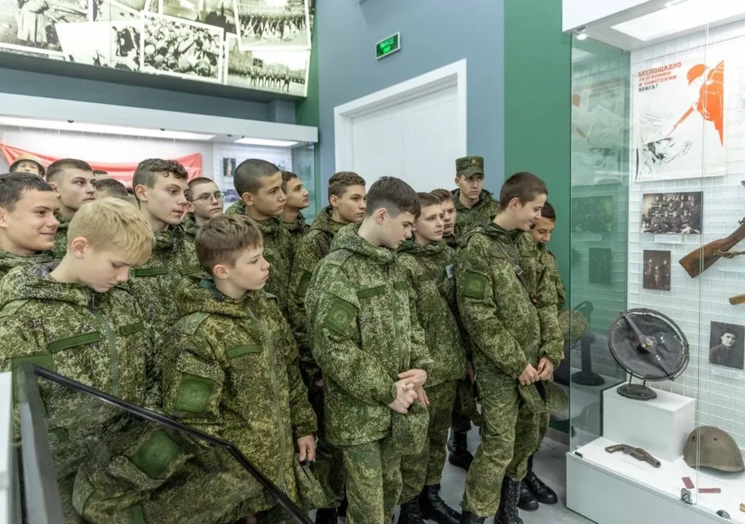 Children and teenagers in matching green camouflage stand beside a museum display of rifles, helmets, and historical photographs.