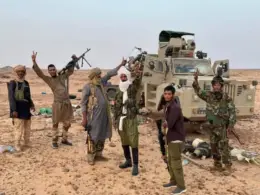 Six armed Tuareg fighters pose beside an armored vehicle in the Malian desert, several raising victory signs.