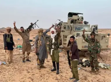 Six armed Tuareg fighters pose beside an armored vehicle in the Malian desert, several raising victory signs.
