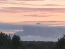 A thick gray plume of smoke rises above a distant tree line beneath a pink early-morning sky.