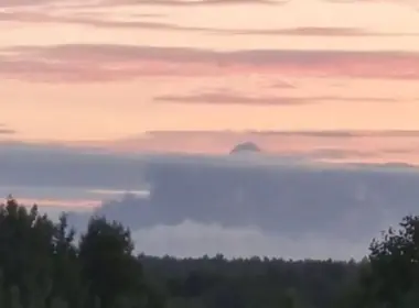 A thick gray plume of smoke rises above a distant tree line beneath a pink early-morning sky.